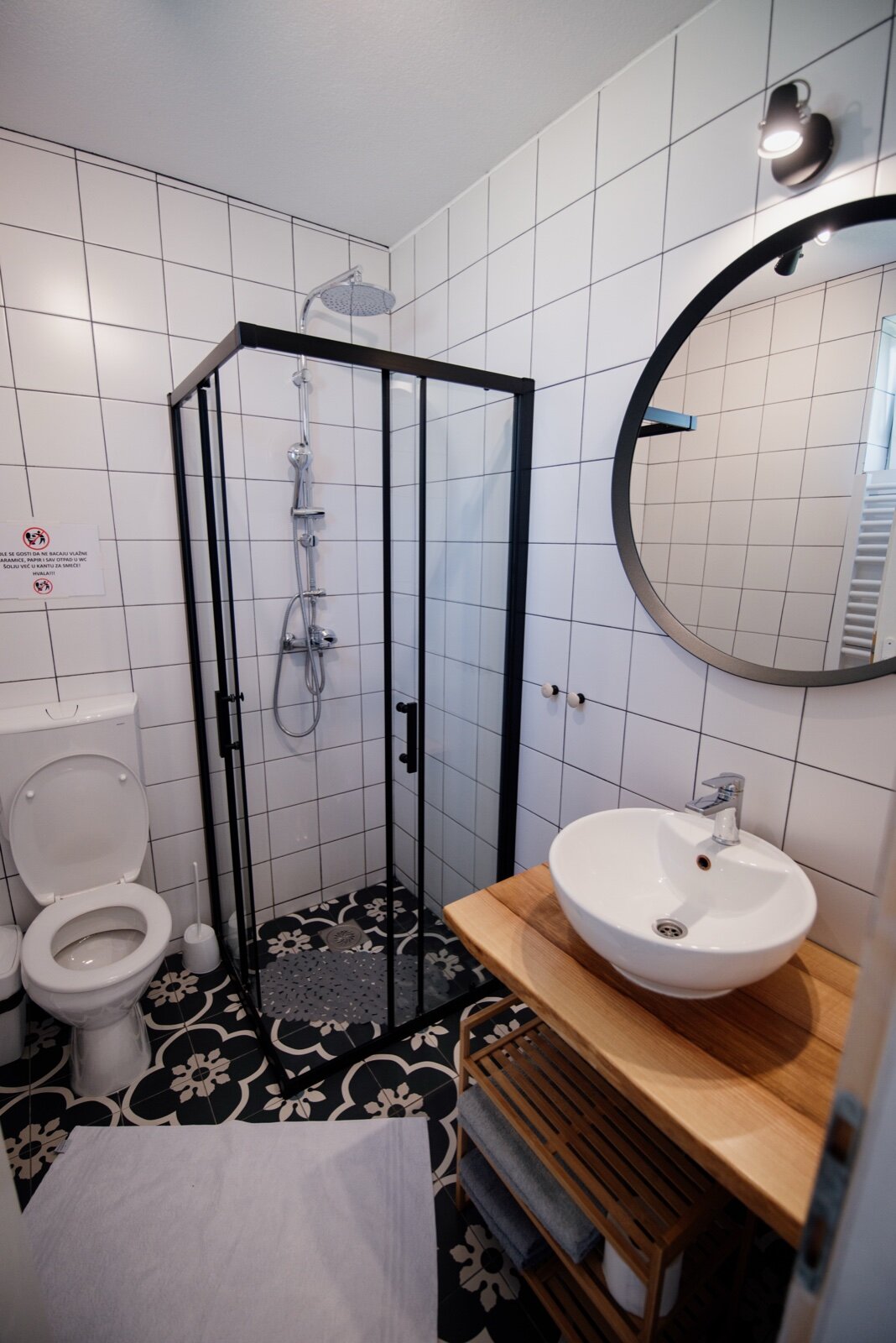 A small modern shower room with a black-framed glass shower enclosure, white square wall tiles, a round mirror, a vessel basin on a timber shelf and a toilet