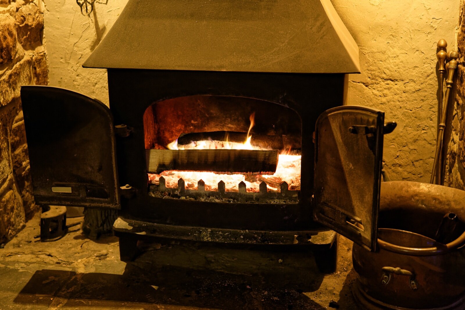 A cast iron wood-burning stove with its doors open, standing on a stone hearth in an inglenook