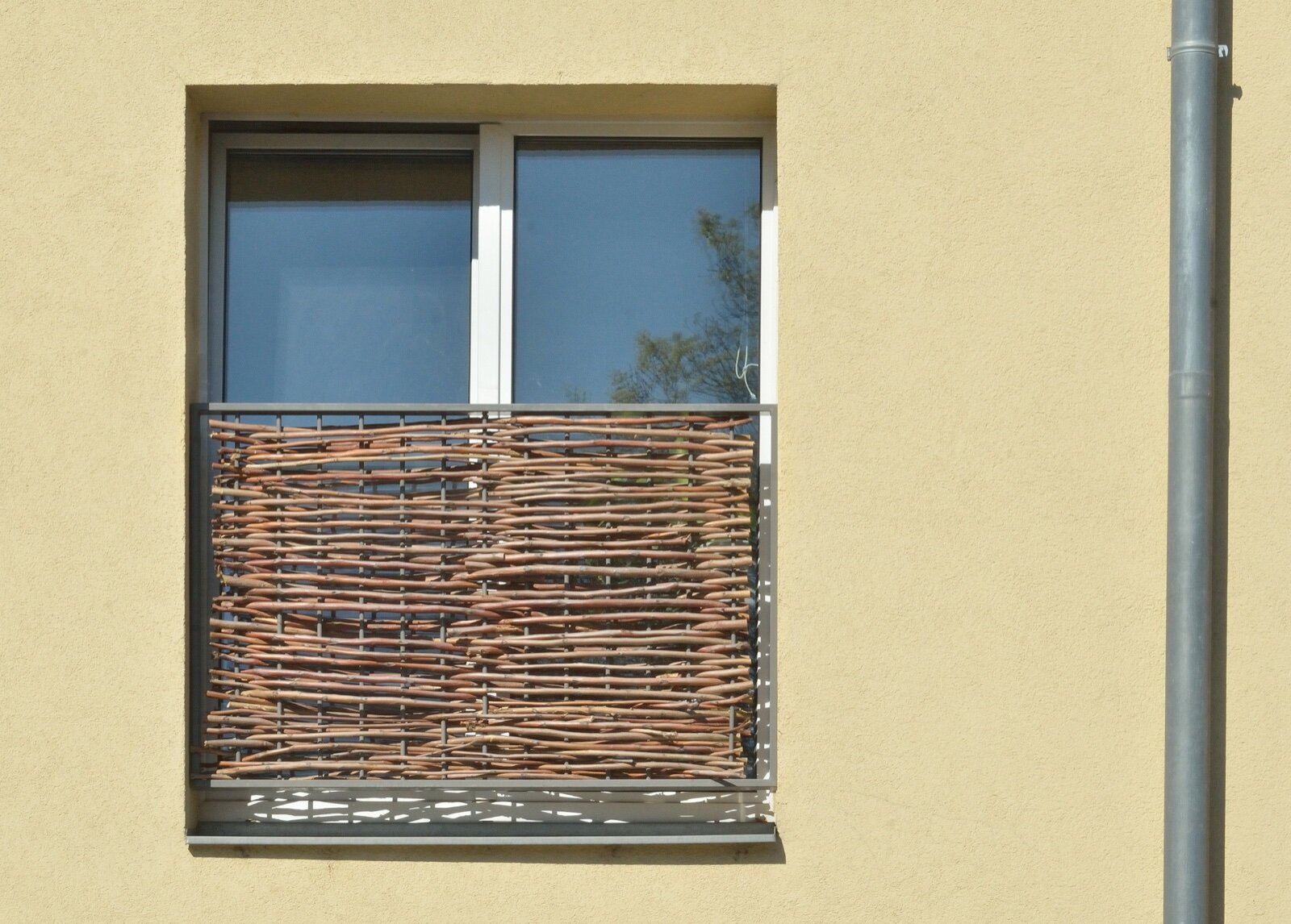 A metal-framed Juliet balcony guard with woven willow infill fixed across a floor-length window on a rendered wall