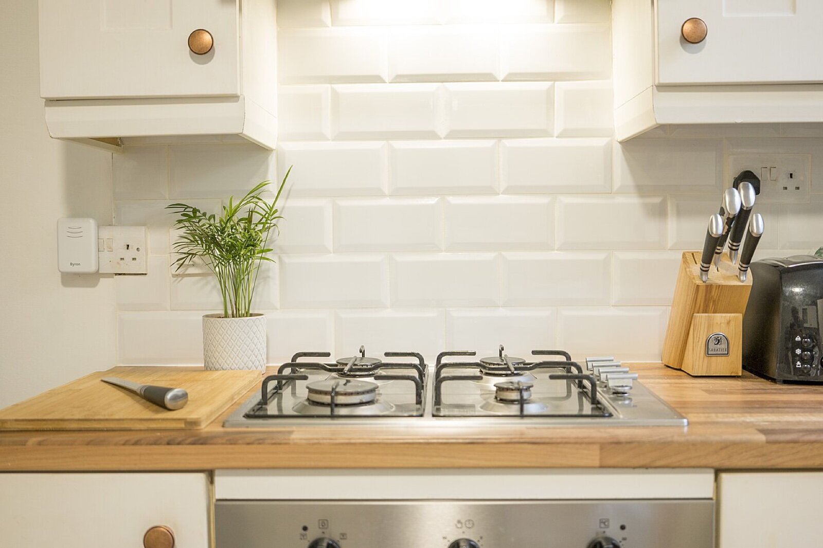 A four-burner gas hob set into a wooden worktop below white metro tiles, with a double socket and a knife block on the counter