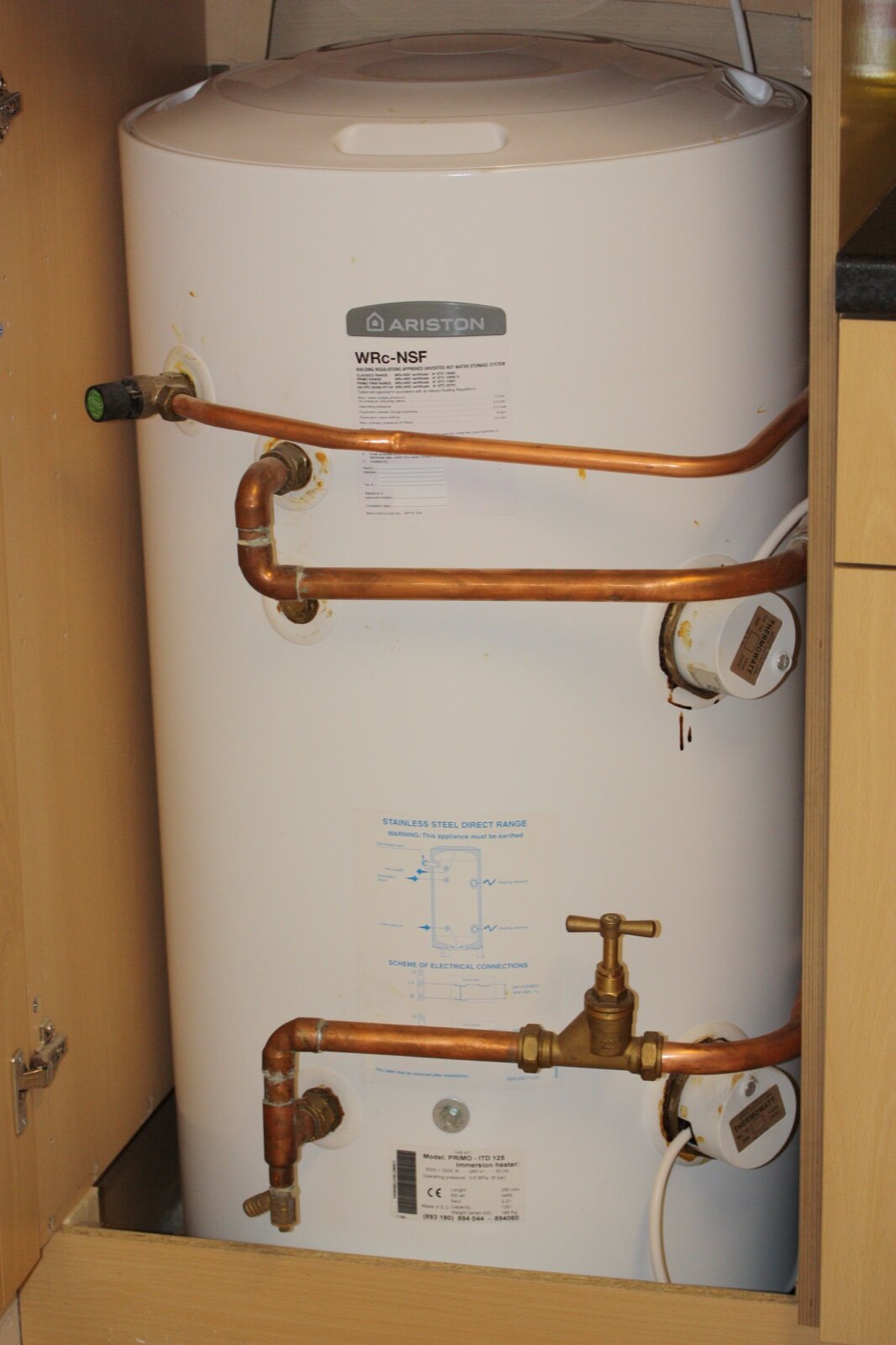 A white unvented hot water cylinder in a fitted cupboard with copper pipework, a pressure relief valve on the side and a building regulations label on the front